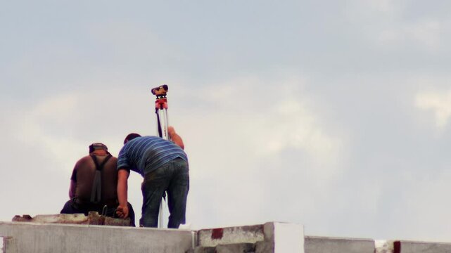 Two male construction workers perform land measurement. One worker adjusts optical leveling instrument tripod on concrete rooftop, other worker sits nearby, looking at horizon. Overcast sky
