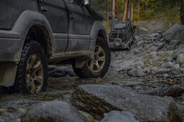 Two off-road vehicles stand in a mountain river within a forest. A medium shot of the right side highlights the aggressive tread of their tires during a summer river crossing.