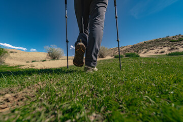 Low-angle close-up of a person's hiking shoes walking on green grass using trekking poles. Sandy hills and clear blue sky in a bright scene.