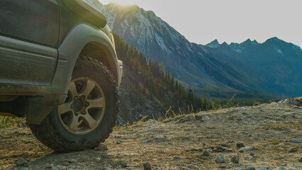 An off-road vehicle is parked on the edge of a stone mountain road. An extreme close-up of its right front tire's aggressive tread is set against a vast autumn valley, forest, and peaks.