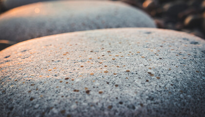 Close-up detail of a smooth, grey pebble or rock surface with tiny brown and white specks, illuminated by a warm, soft light creating subtle highlights and a shallow depth of field.