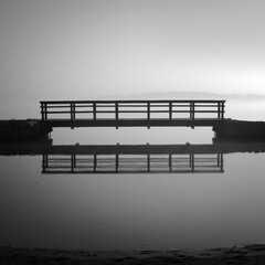 A bridge stretches over still waters its silhouette cast boldly against the calm surface. Black and white art
