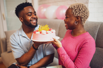 Happy young couple celebrating woman's birthday at home. Boyfriend is giving cake to his girlfriend.