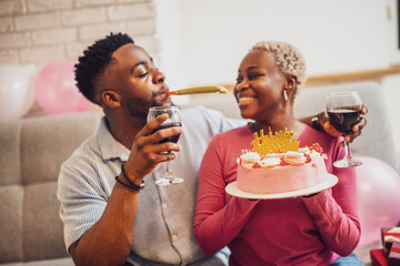 Happy young couple celebrating woman's birthday at home. Woman is holding cake and they are toasting with wine.