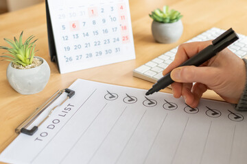 Hand holding marker checking off tasks on a to-do list on a wooden desk, with calendar and plant in background, illustrating productivity, planning, organization and goal progress