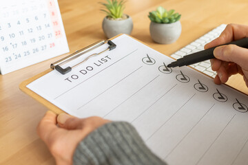 Person's hand marking completed tasks with a black pen on a clipboard, managing workflow and tracking progress, with a calendar and small plants in the background