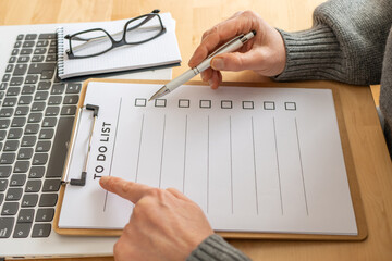 Hands holding a pen pointing to a checklist on a clipboard, marking completed tasks while organizing plans and staying productive at a desk with laptop and glasses