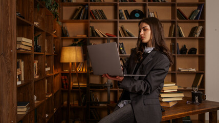 Young woman in a suit jacket holding a laptop in a library with bookshelves and warm lighting © shine.graphics