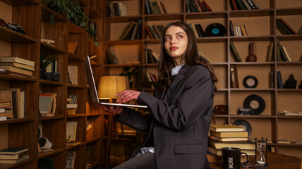 Young woman in a blazer working on a laptop in a cozy library with bookshelves © shine.graphics
