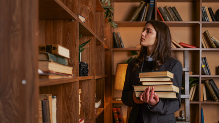 Young woman in a suit holding a stack of books in a library with wooden shelves