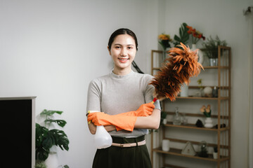 An Asian woman wearing orange rubber gloves smiles while holding a feather duster and spray bottle...