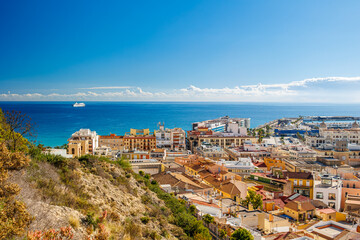 Fototapeta premium Coastal city panorama with sea and harbor view, Panoramic view of a Mediterranean coastal city with colorful rooftops, harbor, hillside foreground, and deep blue sea under clear sky.