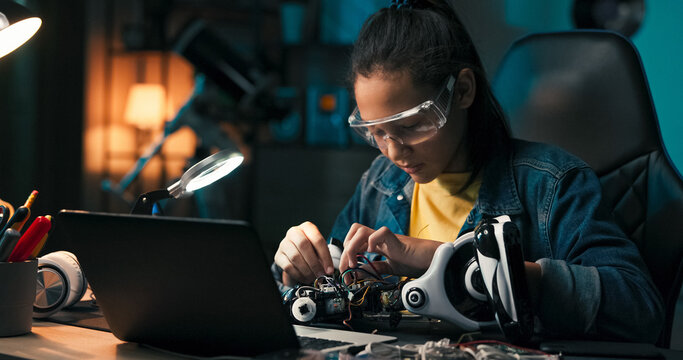 A focused girl builds a robot in a dimly lit room, illuminated only by an office lamp. She connects wires to create a working electronic system, crafting the toy from scratch.
