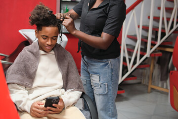 Smiling afro young man checking phone while stylist braids his hair from the side