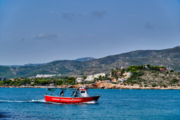 Red fishing boat sails near coastal landscape with hills and houses under clear blue sky