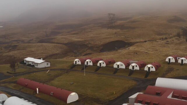 Aerial drone view of red and white Quonset huts aligned on green lawns in Hvalfj&ouml;r&eth;ur Iceland with foggy hills and rural landscape in the background during overcast weather