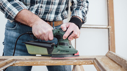 Craftsman. Adult carpenter using an electric sander to smooth an old wooden window. Construction industry, carpentry, housework do it yourself. Restoration.