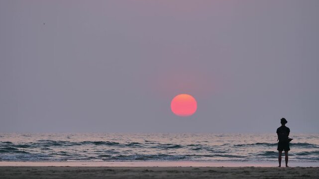 man standing on quiet beach watching large pink sun setting over calm ocean creating reflective mood
