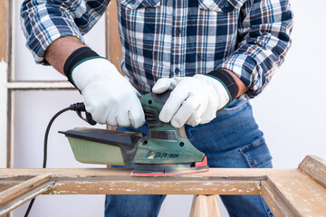 Craftsman. Adult carpenter using an electric sander to smooth an old wooden window. Construction industry, carpentry, housework do it yourself, workplace safety. Restoration.