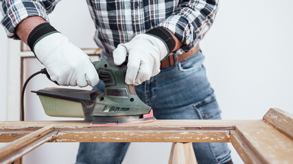 Craftsman. Adult carpenter using an electric sander to smooth an old wooden window. Construction industry, carpentry, housework do it yourself, workplace safety. Restoration.