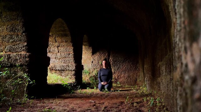 woman kneeling in dim ancient stone arch interior with calm expression and soft light creating quiet meditation mood
