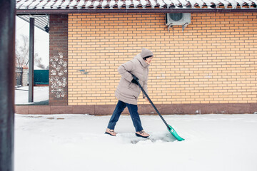 A young Asian woman in a beige winter coat shovels snow outside a brick house, while clearing the snow.