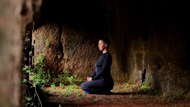 Woman kneeling in dim cave-like space with eyes closed and hands resting together showing calm meditation amid earthy stone walls and soft natural light
