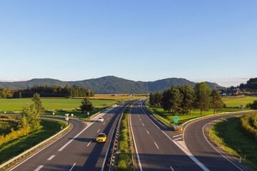 Cars travel on a highway surrounded by green fields and mountains, highlighting mobility and urban development during busy hours.