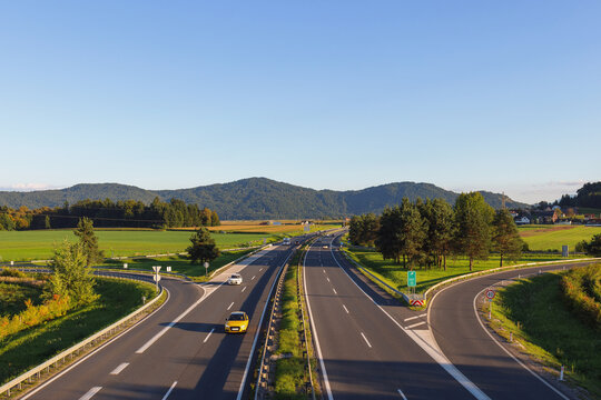 Cars travel on a highway surrounded by green fields and mountains, highlighting mobility and urban development during busy hours.