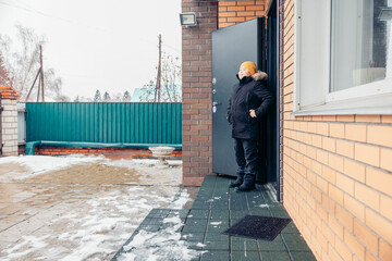 young Asian boy with a yellow hat stands at the door of a brick house during winter. Snow is visible on the ground outside, child safety on the street