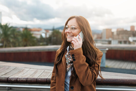 Cheerful woman with long hair and glasses talking on smartphone, wearing brown coat, standing outside in city with cloudy sky and soft light