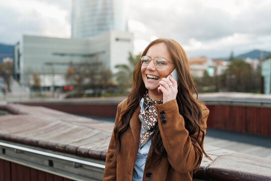Woman wearing brown coat and glasses laughing while talking on phone, urban background with modern buildings, concept of communication