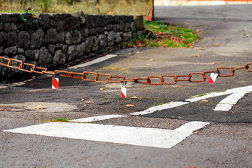 Rusty metal chain blocking an empty roadway. Outdoor barrier with warning reflectors. White road markings on asphalt. Estricted access and closed passage