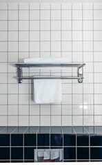 A clean bathroom with white ceramic tiles. A metal towel rack holds two neatly folded white towels. The floor features dark ceramic tiles.
