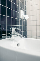 A modern bidet with a chrome faucet in a tiled bathroom. The bidet features a sleek design and is positioned against a backdrop of dark and light tiles.