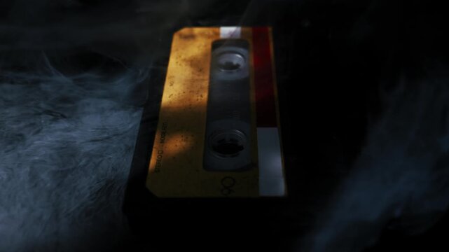 Vintage audio cassette tape with a yellow label lies on a dark reflective surface. Mysterious white smoke billows and swirls, obscuring the retro object.