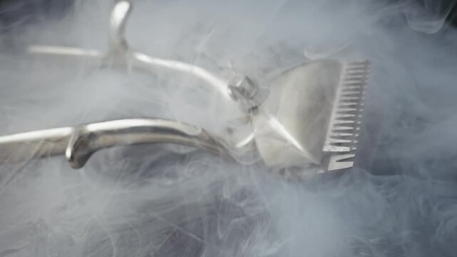 Vintage manual hair clipper rests on a dark surface while white smoke billows around it. Slow-motion camera pans around the metallic tool, creating a dramatic, mysterious mood.