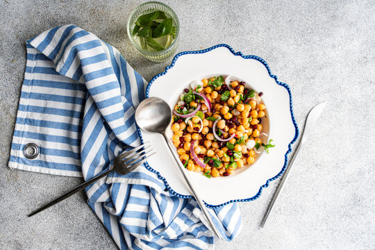 Overhead view of a bowl of chickpea, red onion, pomegranate and coriander salad with a glass of mint water on a table