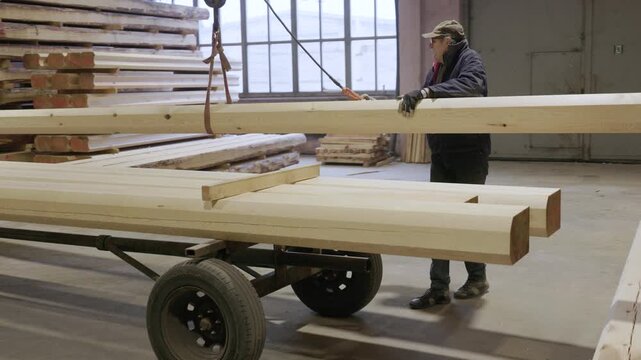 A worker handles a long milled timber beam using a lifting strap in a lumber workshop, placing it onto a trolley stacked with other beams. Natural light from large windows