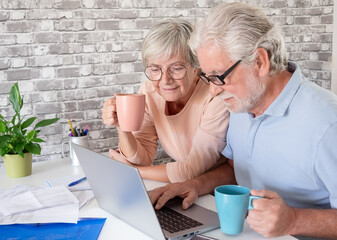 Happy senior couple using a laptop together at home, looking at the screen while managing paperwork. Concept of active aging, digital literacy, online banking, communication, technology