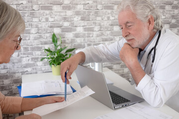 Senior male doctor consulting with elderly female patient in a medical office, reviewing documents at a desk with a laptop. Concept of healthcare, medical consultation, diagnosis, trust, patient care