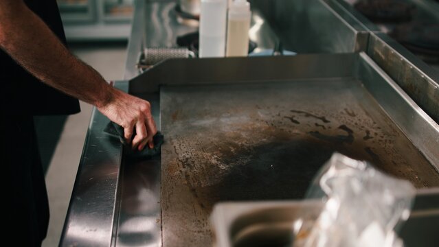 Chef cleaning a flat top griddle in a dark commercial kitchen for food preparation