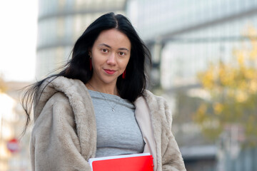 A young woman holds a red notebook and confidently looks at the camera on the street.