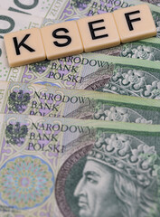 Blocks with the abbreviation KSEF, standing for National E-invoice System, are placed against a background of loosely spread Polish 100 PLN banknotes. The photo was taken under artificial lighting.