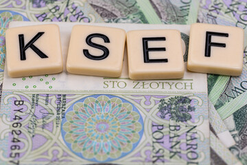 Blocks with the abbreviation KSEF, standing for National E-invoice System, are placed against a background of loosely spread Polish 100 PLN banknotes. The photo was taken under artificial lighting.