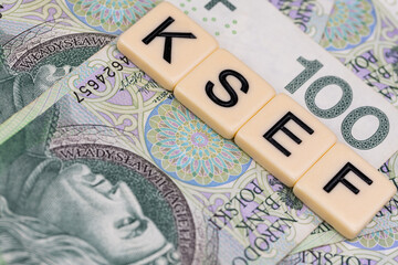 Blocks with the abbreviation KSEF, standing for National E-invoice System, are placed against a background of loosely spread Polish 100 PLN banknotes. The photo was taken under artificial lighting.