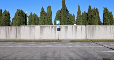 Blue parking sign in front of a vacant car park. Long surrounding concrete wall, cypresses and sky on behind. Background for copy space.