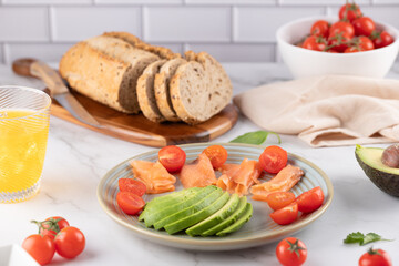 Healthy breakfast with whole grain bread, salmon, avocado and cherry tomatoes. White bowl and orange kombucha glass. Light background. Concept of balanced food.