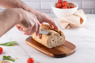Man's hands slicing fresh whole grain bread on a wooden board, on marble countertop. Cherry tomatoes and basil in the background. Tiled and marble setting. 