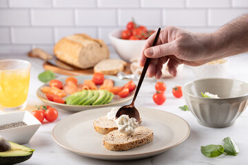 Male hand adding fresh cheese to whole grain toasts on ceramic plate. Healthy ingredients in the background. Concept of mindful eating and healthy cooking